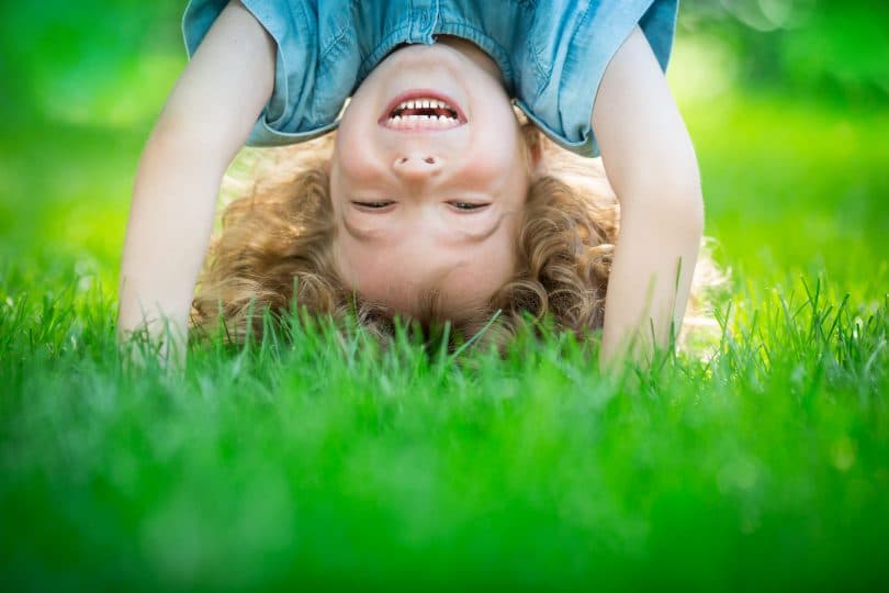 Uma criança sorridente plantando bananeira em um gramado verde. A criança é um menino de cabelos cacheados loiros.