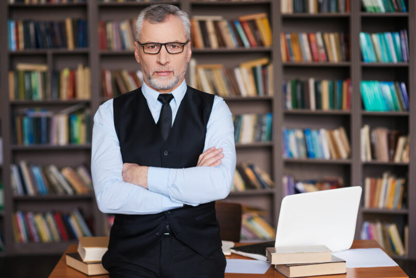 Professor em pé em biblioteca. Ele se apoia em uma mesa cheia de livros. Usa óculos e tem cabelo grisalho.