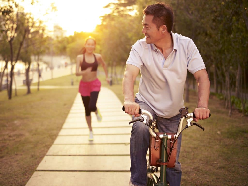 Casal asiático em um parque. A mulher está ao fundo correndo e o homem está a frente andando de bicicleta.