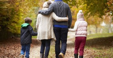 Família andando de mãos dadas em um caminho de folhas. Um menino. Uma menina. Um homem. Uma mulher. Todos usam roupas de frio.