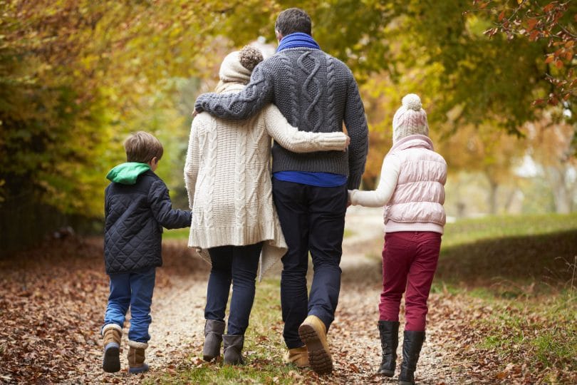 Família andando de mãos dadas em um caminho de folhas. Um menino. Uma menina. Um homem. Uma mulher. Todos usam roupas de frio.