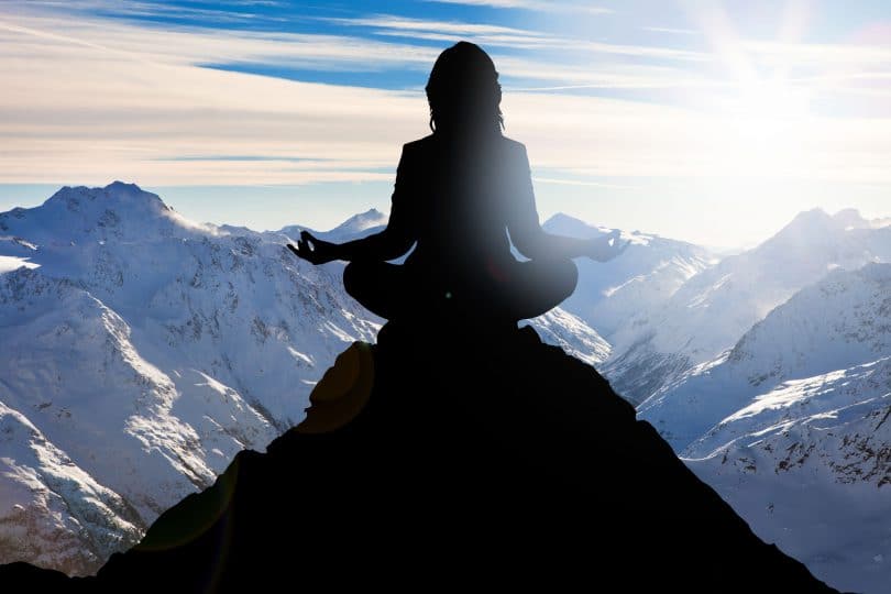 Silhueta de uma mulher sentada no pico de uma montanha meditando.
