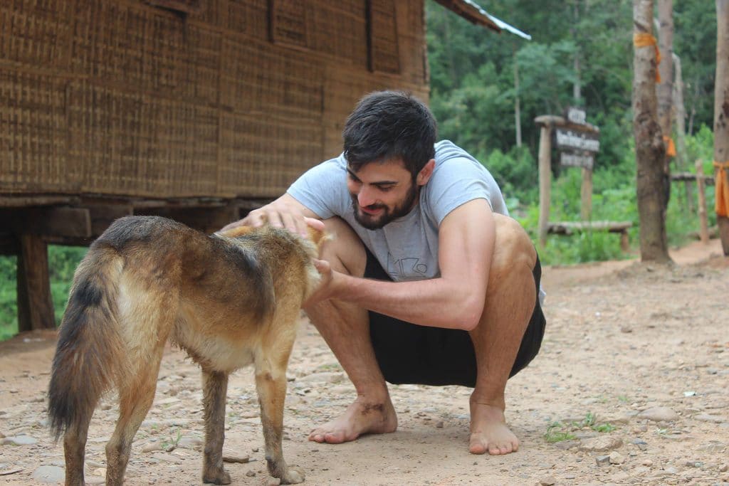 Rapaz branco de cabelos pretos e barbado agachado e fazendo carinho em um cachorro.