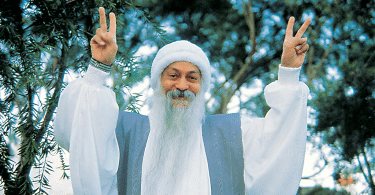 Osho, vestido de branco, fazendo o sinal da paz com as duas mãos, com árvores ao fundo.