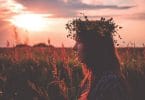 Menina branca, usando uma coroa de flores, no meio de um campo de flores, com um pôr do sol ao fundo.