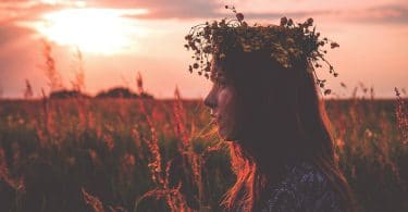 Menina branca, usando uma coroa de flores, no meio de um campo de flores, com um pôr do sol ao fundo.