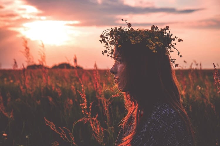 Menina branca, usando uma coroa de flores, no meio de um campo de flores, com um pôr do sol ao fundo.