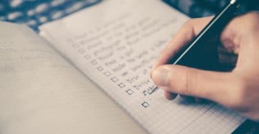 Mão de pessoa branca escrevendo em uma agenda uma lista de tarefas.