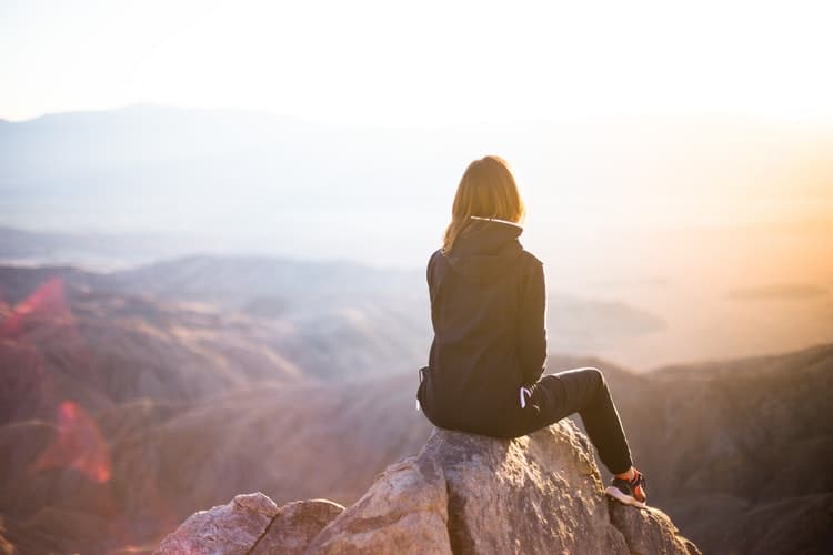 Mulher sentada em topo de montanha, observando o pôr do sol.