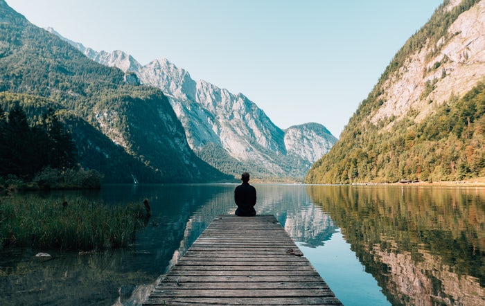 Homem vestido de preto, sentado em pier, observando lago, montanhas e céu azul.