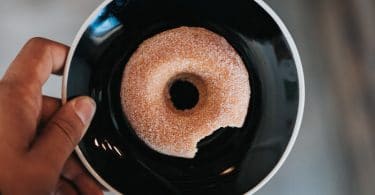 Mão de homem negro segurando um prato preto pequeno, com uma rosquinha açucarada mordida.