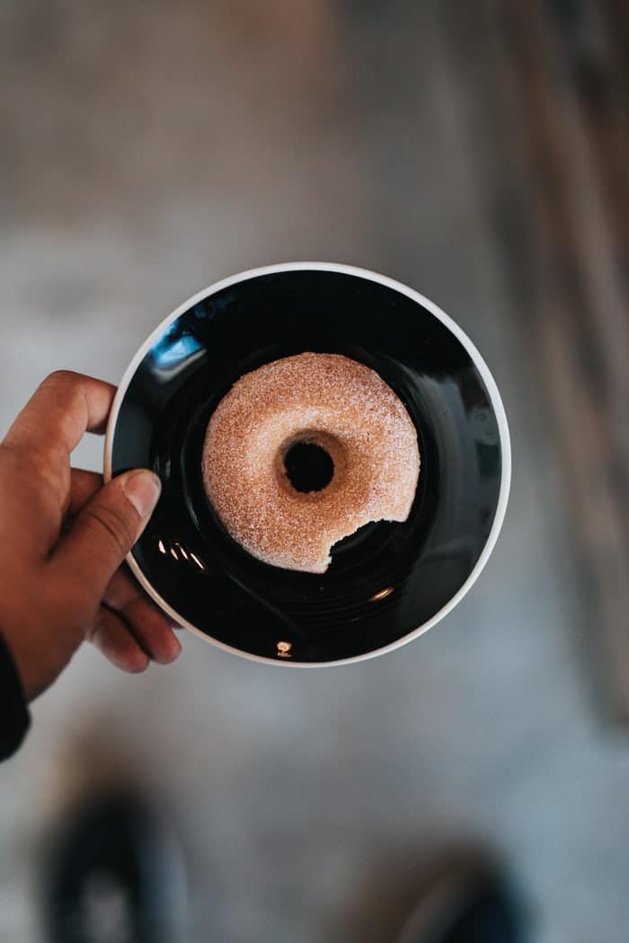 Mão de homem negro segurando um prato preto pequeno, com uma rosquinha açucarada mordida.