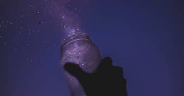 Mão de homem branco segurando pote de vidro para cima, apontando para o céu noturno estrelado.