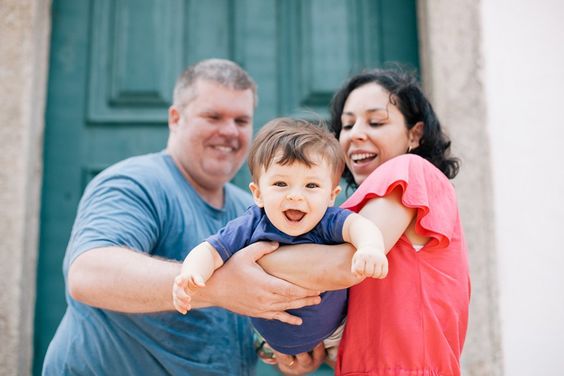 Família composta por mãe, pai e filho. Os pais seguram o filho como se estivesse voando. A mãe é morena e usa uma blusa vermelha. O pai é grisalho e usa uma blusa azul. O filho é moreno e tem o cabelo de tigela e também usa uma blusa azul.