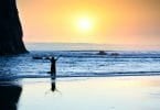 Silhueta de menina em pé nas ondas da praia com os braços levantados ao pôr do sol.
