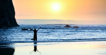 Silhueta de menina em pé nas ondas da praia com os braços levantados ao pôr do sol.