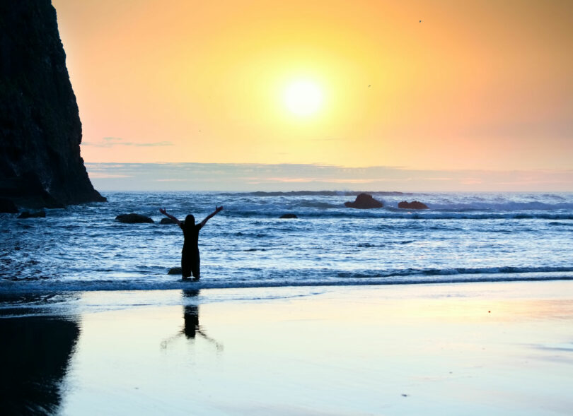 Silhueta de menina em pé nas ondas da praia com os braços levantados ao pôr do sol.