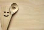 Duas colhes de pau de cozinha, uma com um coração e uma com um rosto sorridente entalhadas, ambas em cima de uma mesa de madeira.