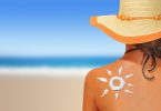 Mulher na praia usando chapéu de palha de costas para a foto. Em suas costas há um desenho feito com protetor solar em formato de sol.