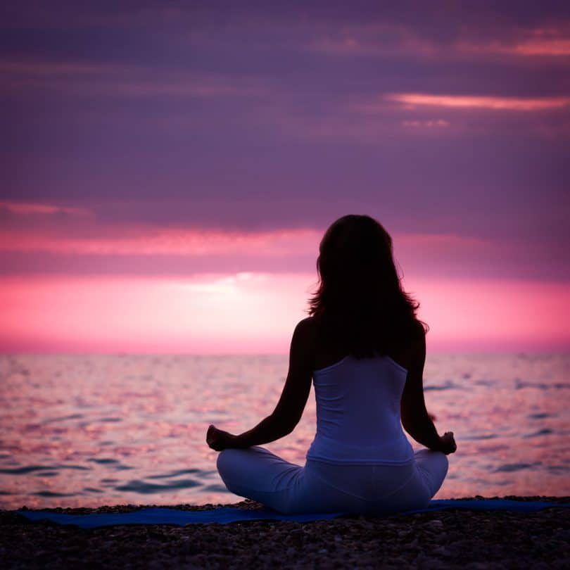 Silhueta da mulher que medita na posição de lótus pelo mar no por do sol.