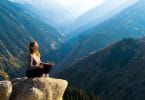 Mulher meditando no alto de uma montanha. Ao fundo uma paisagem de céu azul e montanhas.