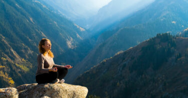 Mulher meditando no alto de uma montanha. Ao fundo uma paisagem de céu azul e montanhas.