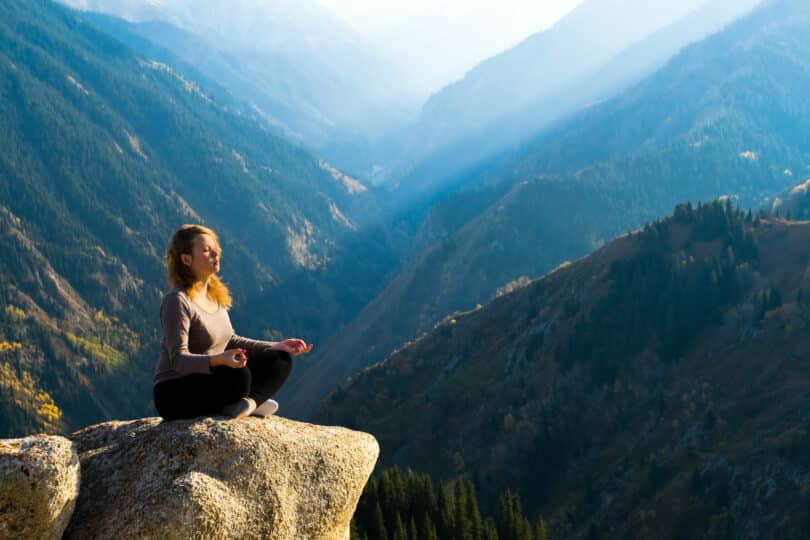 Mulher meditando no alto de uma montanha. Ao fundo uma paisagem de céu azul e montanhas.