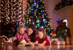 Mãe com dois filhos pequenos sentados no meio da sala com decorações de natal, lendo um livro com as crianças.