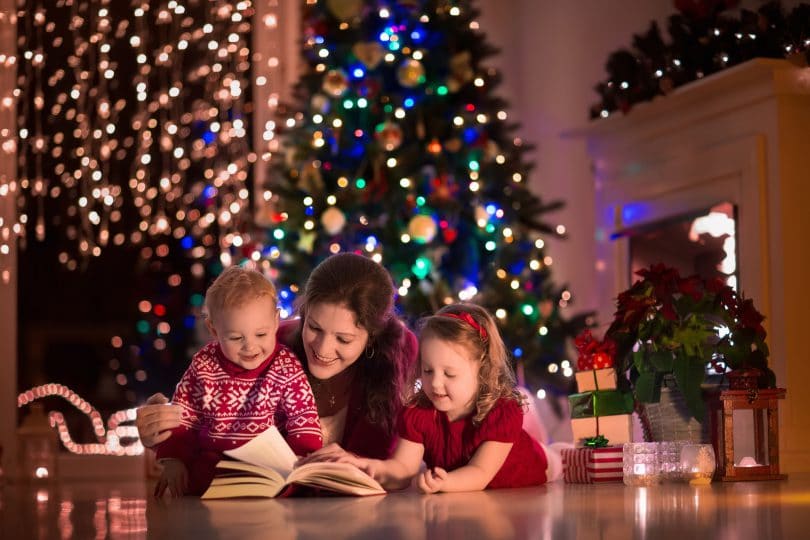 Mãe com dois filhos pequenos sentados no meio da sala com decorações de natal, lendo um livro com as crianças.