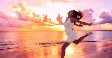 Mulher jovem e sorridente, correndo na praia, a beira mar, com o por do sol ao fundo.