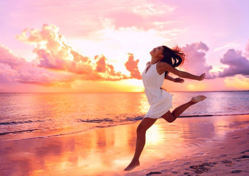 Mulher jovem e sorridente, correndo na praia, a beira mar, com o por do sol ao fundo.