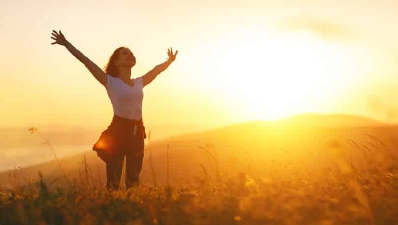 Mulher em pé de braços abertos com sol ao fundo