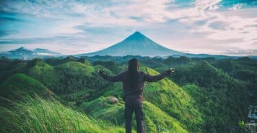 Homem, com os braços abertos em cima de uma montanha com vista para outras montanhas verdes