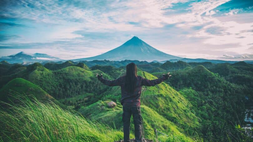 Homem, com os braços abertos em cima de uma montanha com vista para outras montanhas verdes