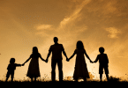 Silhueta de família, com homem, mulher e três crianças, de mãos dadas olhando o pôr do sol.