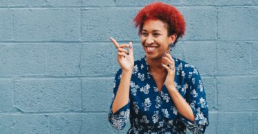 felicidade. Mulher negra de cabelos encaracolados e vermelhos. Ela está sorrindo e apontado para o lado.