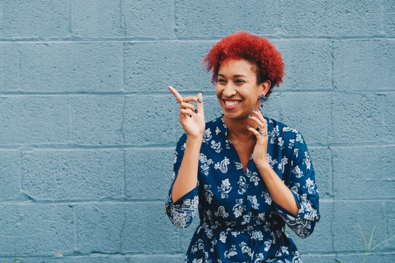 felicidade. Mulher negra de cabelos encaracolados e vermelhos. Ela está sorrindo e apontado para o lado.