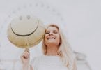 Mulher sorridente em um parque de diversões, posando para foto em frente de uma roda gigante enquanto segura um balão de carinha sorridente.