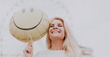 Mulher sorridente em um parque de diversões, posando para foto em frente de uma roda gigante enquanto segura um balão de carinha sorridente.