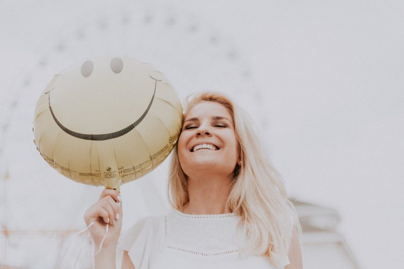 Mulher sorridente em um parque de diversões, posando para foto em frente de uma roda gigante enquanto segura um balão de carinha sorridente.