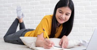 Adolescente asiática deitada na cama enquanto faz anotações em um caderno em frente ao computador.