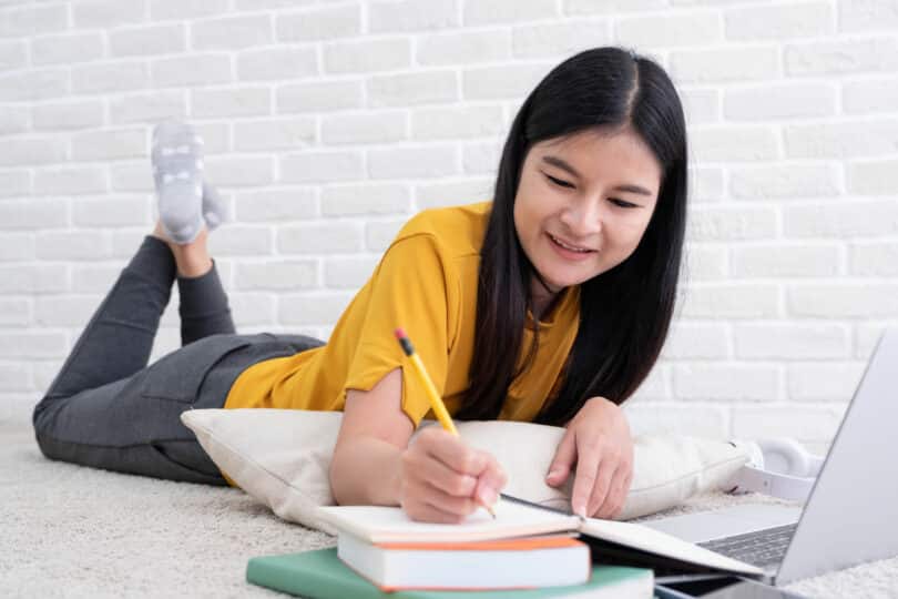 Adolescente asiática deitada na cama enquanto faz anotações em um caderno em frente ao computador.