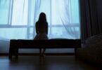 Menina sentada em uma cama olhando pela janela.