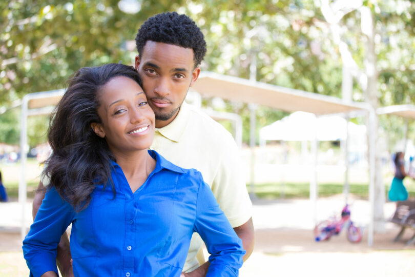 Casal hétero negro posando para foto abraçados com paisagem de parque ao fundo.