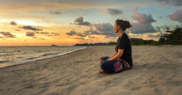 Mulher em posição meditativa sentada na areia.