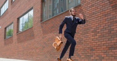 Homem vestido como executivo, de terno e pasta. Ele corre em frente à uma parede de tijolos.