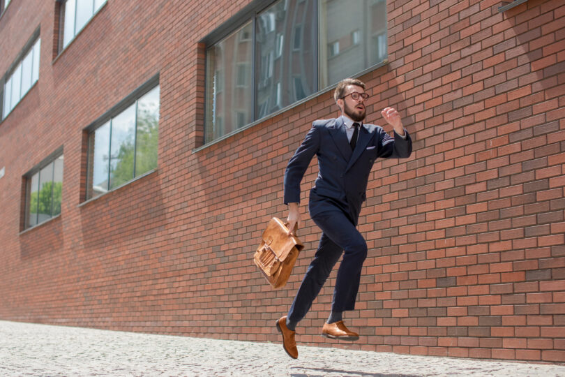 Homem vestido como executivo, de terno e pasta. Ele corre em frente à uma parede de tijolos.