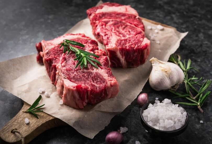 Dois bifes de carne vermelha postos em cima de papel vegetal e uma tábua de madeira. Sal grosso, alho e alecrim ao lado.