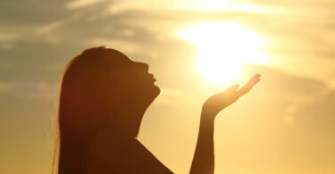 Silhueta de mulher com a mão estendida para a luz que emana do sol.