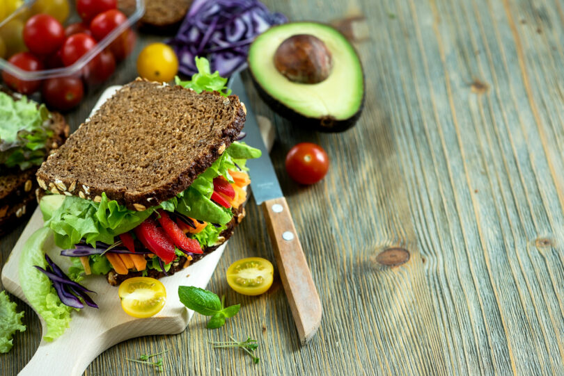 Sanduíche vegano com ingredientes frescos: abacate, alface, tomate, cenoura, repolho roxo e uma faca.